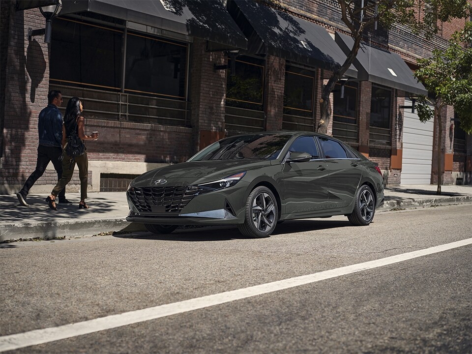 Hyundai Elantra hybrid parked in the city, with a couple walking on the sidewalk, illustrating an urban and modern lifestyle.