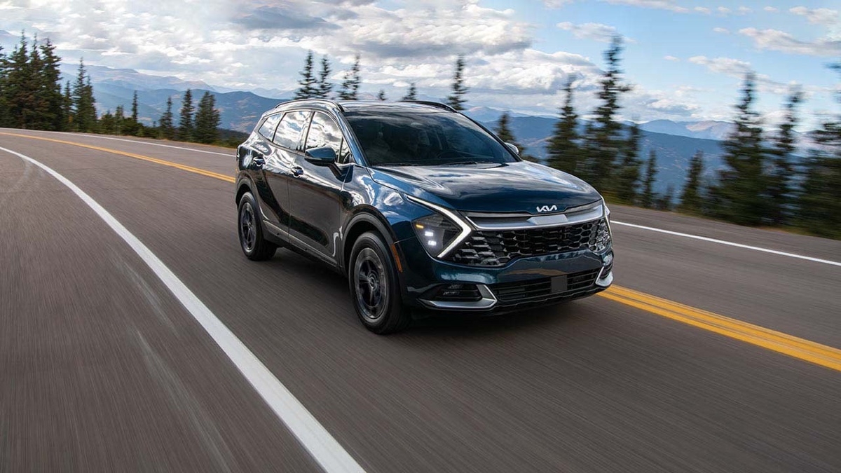 Black Kia Sportage hybrid driving on a mountain road lined with trees, with a cloudy sky in the background.