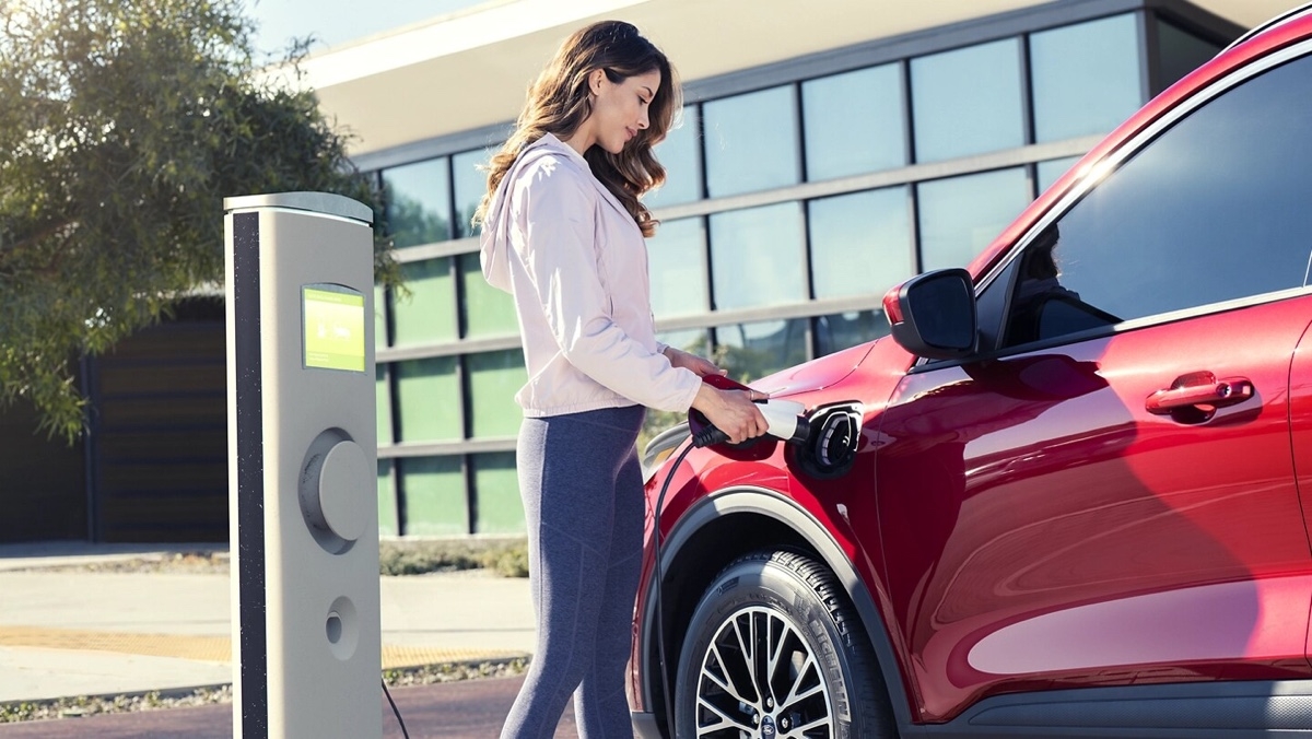 Woman charging a red hybrid SUV at a charging station in front of a modern building, illustrating a thoughtful step in the purchase of an electrified vehicle.